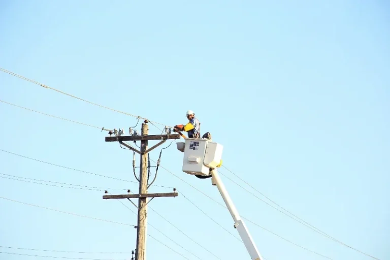 A worker in a cherry picker repairs a utility pole against a clear sky.
