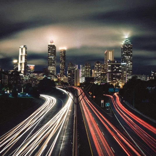 City skyline at night with illuminated buildings and streaks of car lights on a highway.
