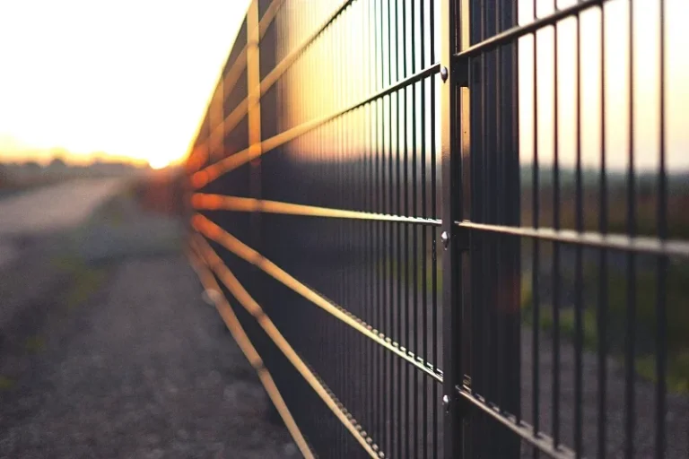 Boundary fencing Close-up of a metal fence with a sunset in the background, casting reflections.