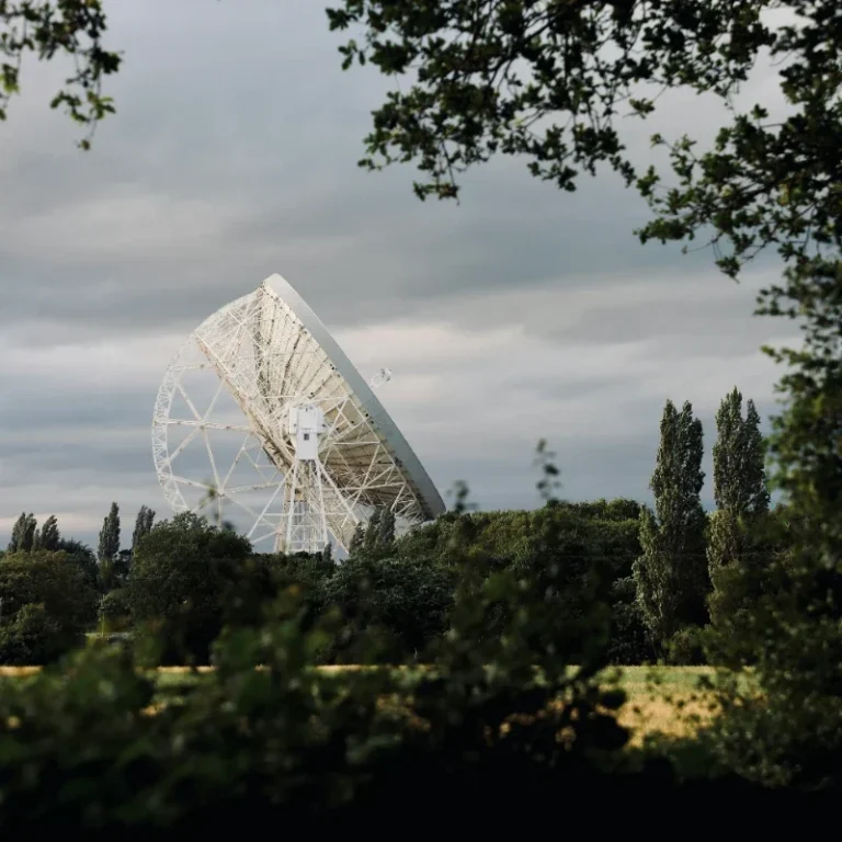 A large radio telescope, Jodrell Bank, stands against a cloudy sky, surrounded by trees.